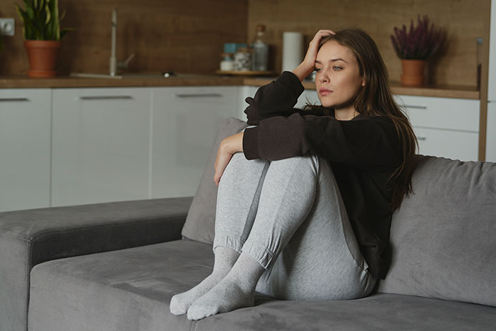 Young woman sitting on couch looking thoughtful, contemplating sister’s $4k plastic surgery and body dysmorphia concerns. Young woman sitting on couch looking thoughtful, contemplating sister’s $4k plastic surgery and body dysmorphia concerns.