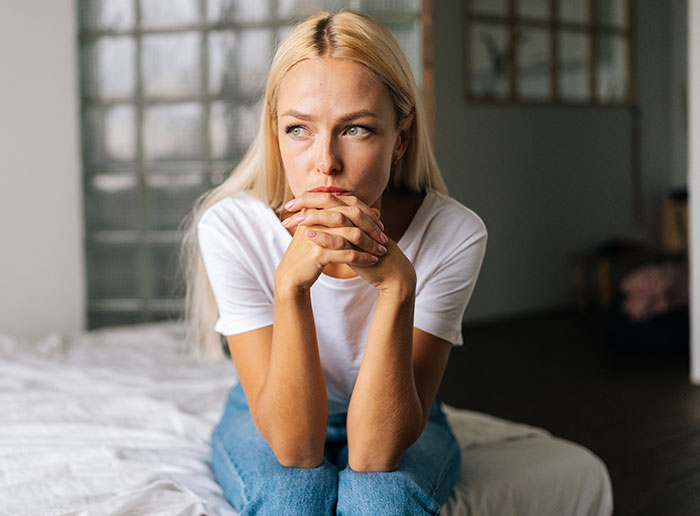 Woman refusing homeless sister-in-law to move in, sitting thoughtfully on bed in a casual white shirt and jeans. Woman refusing homeless sister-in-law to move in, sitting thoughtfully on bed in a casual white shirt and jeans.