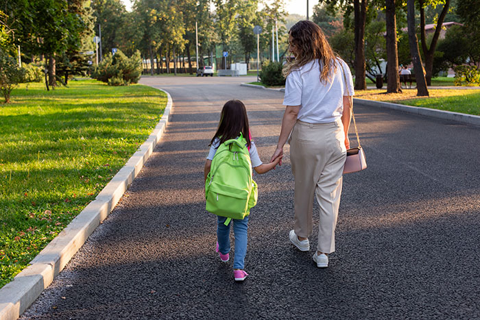 Woman holding niece's hand walking to school on quiet road surrounded by trees in early morning light Woman holding niece's hand walking to school on quiet road surrounded by trees in early morning light