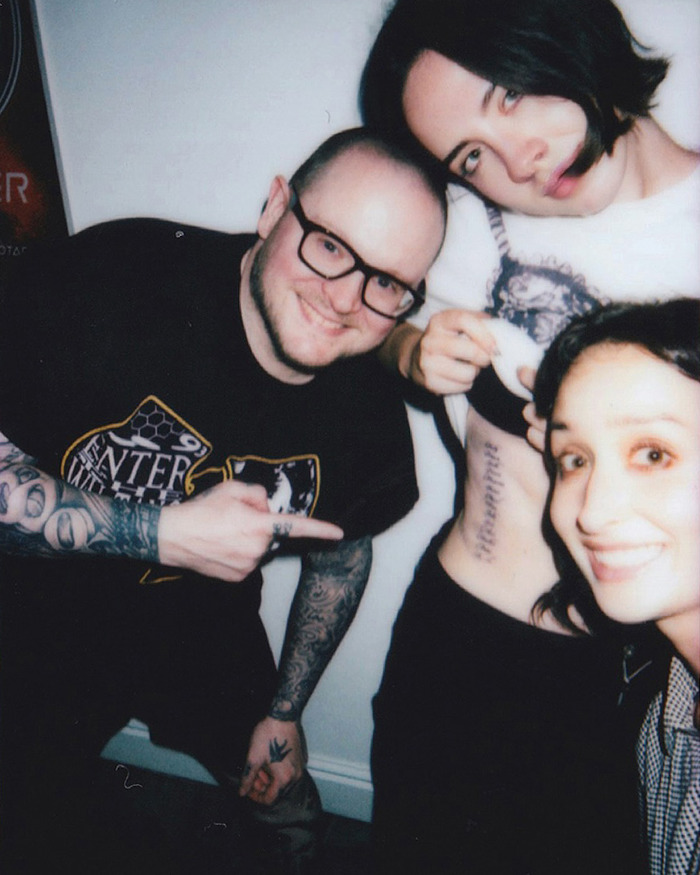 Three people in a casual setting showing a woman's feminist tattoo on her side, sparking internet debate and controversy.