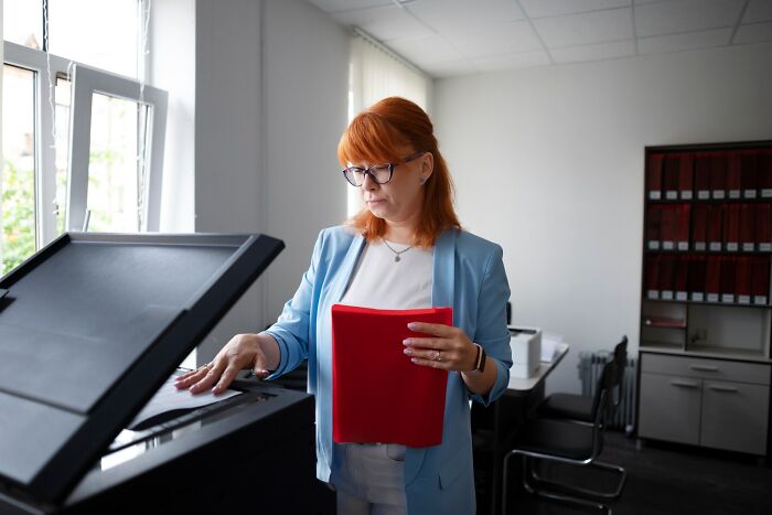 Woman in glasses using office copier, holding red folder, illustrating infuriating work habits at office environment.