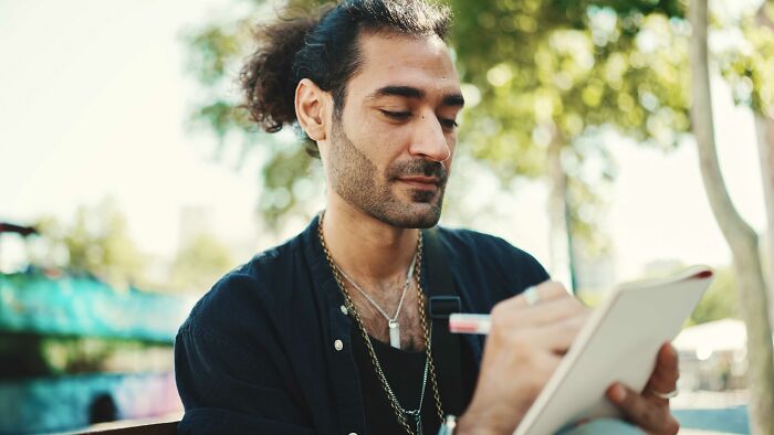 Man with curly hair writing in a notebook outdoors, capturing real-life horror story moments in a peaceful setting.