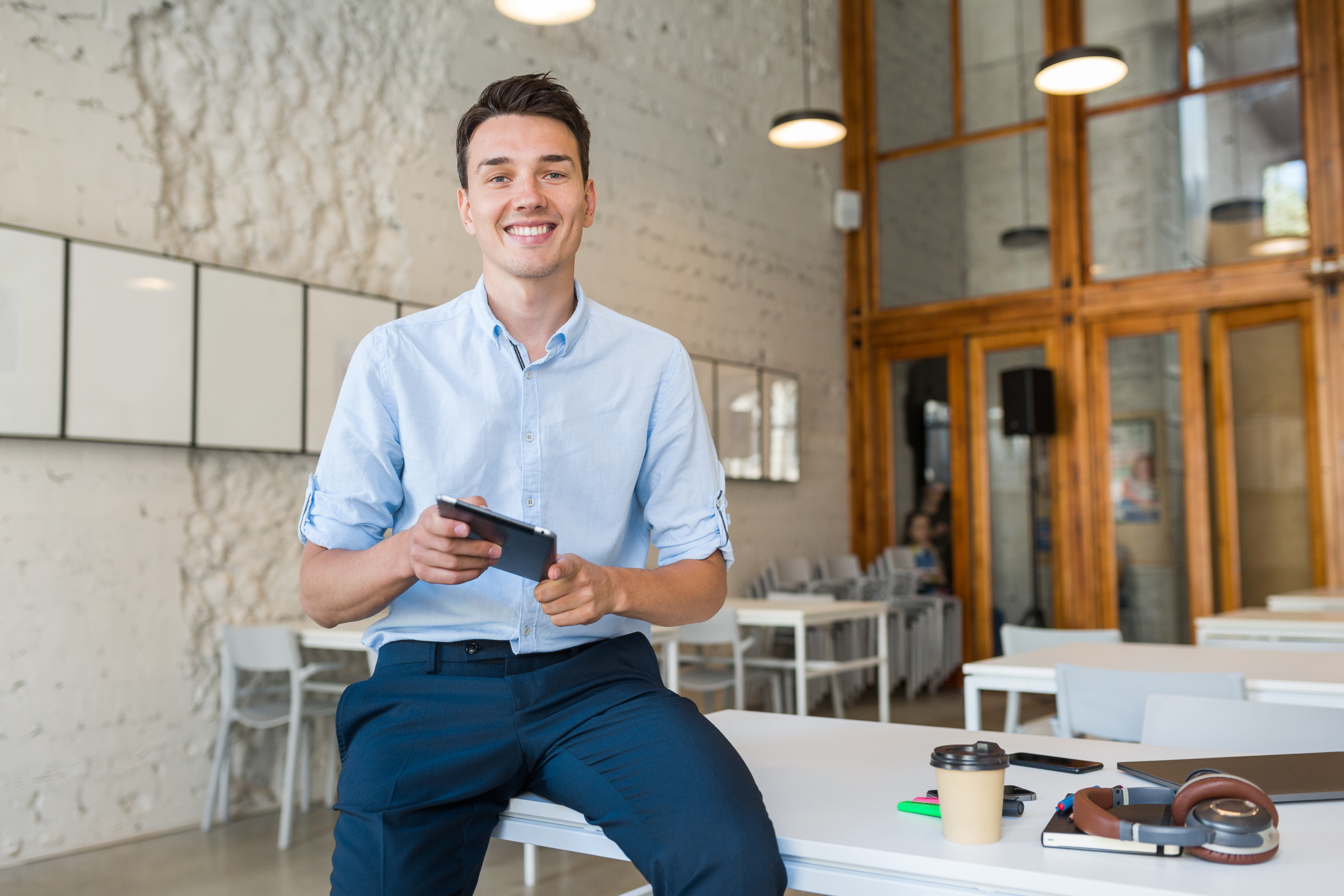 Young man in smart casual attire smiling while holding a phone in a modern office, capturing corporate comedy gold vibe.