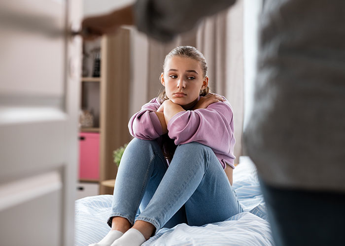 Teen girl sitting on bed looking upset while dad stands in doorway, highlighting babysitting step-siblings conflict.