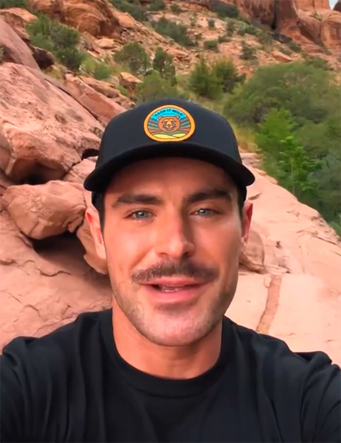 Zac Efron outdoors wearing a black cap, with a natural rocky background, showing his face in a casual selfie. Zac Efron outdoors wearing a black cap, with a natural rocky background, showing his face in a casual selfie.