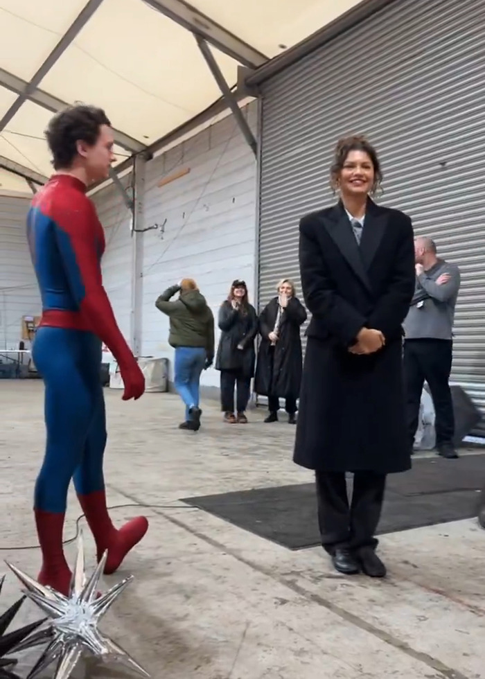 Zendaya smiling and standing near Tom Holland in Spider-Man costume on a set with several people in the background.