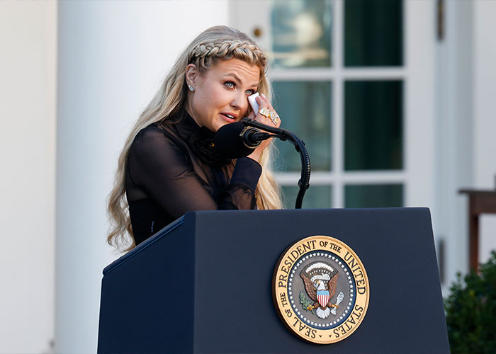 Erika Kirk wiping a tear while speaking behind a presidential podium in an emotional outdoor setting. Erika Kirk wiping a tear while speaking behind a presidential podium in an emotional outdoor setting.