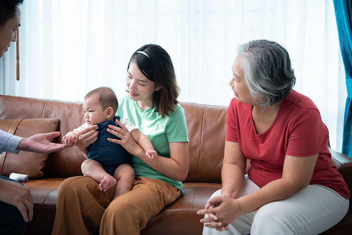 Mother-in-law expressing concern to daughter-in-law holding premature baby while sitting with family on a couch.