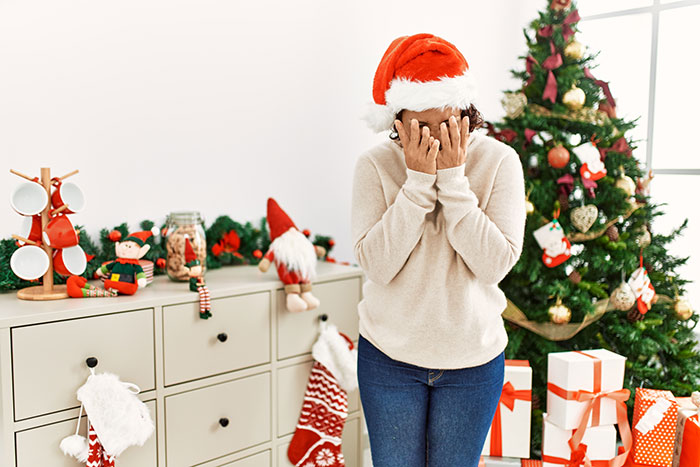 Woman in Santa hat covering face next to Christmas tree and gifts, reflecting on deserving another Christmas gift reality check. Woman in Santa hat covering face next to Christmas tree and gifts, reflecting on deserving another Christmas gift reality check.