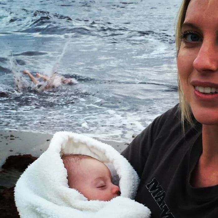 Woman holding a baby wrapped in a blanket on the beach with someone splashing in the water captured at the perfect time