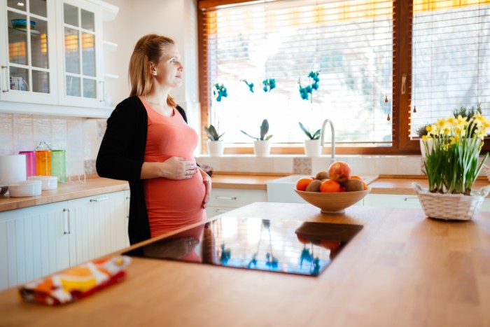 Pregnant woman standing in a bright kitchen, reflecting on family issues as husband won’t stand up to his mother.