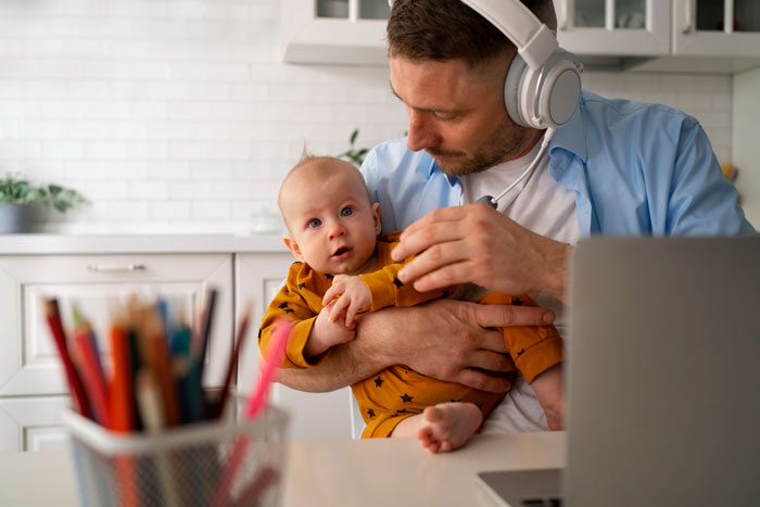 Man wearing headphones holding baby in a cozy home office, symbolizing a new mom hiding from religious family conflict.