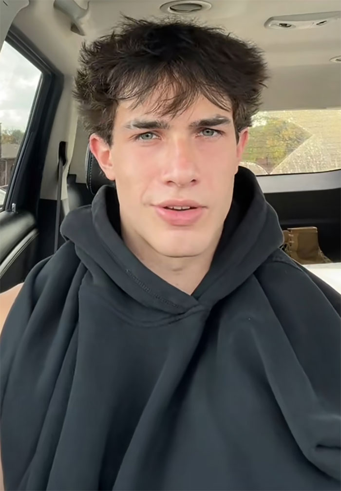 Young man with messy hair wearing a black hoodie inside a car, sharing a story about a waiter serving first date disaster.