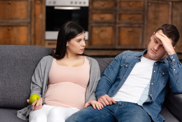Pregnant woman looks frustrated while husband won’t stand up to his mother, causing tension in their relationship at home.