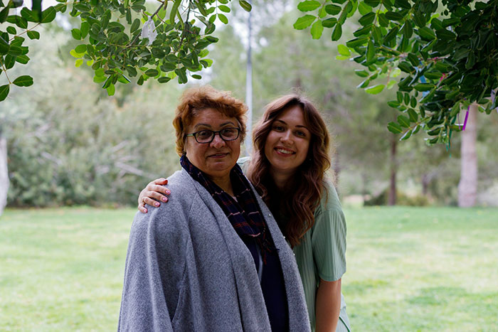 Middle-aged woman with glasses and younger woman smiling outdoors under leafy trees, reflecting husband and mother tension.