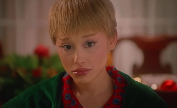 Ariana Grande in SNL Home Alone parody wearing short blonde wig and floral red shirt with green robe in a festive setting.