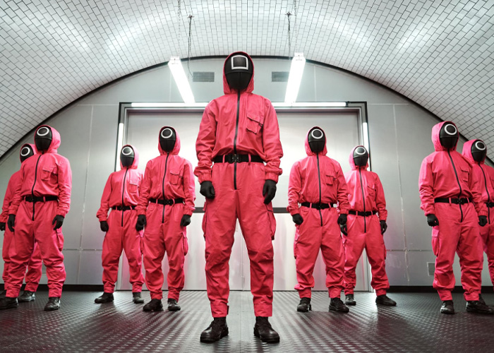 Group of people in red costumes and black masks resembling Squid Game characters in a dimly lit room setting. Group of people in red costumes and black masks resembling Squid Game characters in a dimly lit room setting.