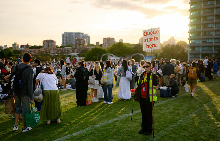 Crowd gathered on grassy public space as a staff member holds a sign marking the start of a queue on New Year&rsquo;s Eve.
