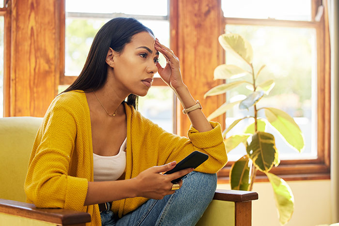 Woman sitting indoors looking stressed and holding a phone during a difficult wedding dress shopping trip and friendship wake-up call.