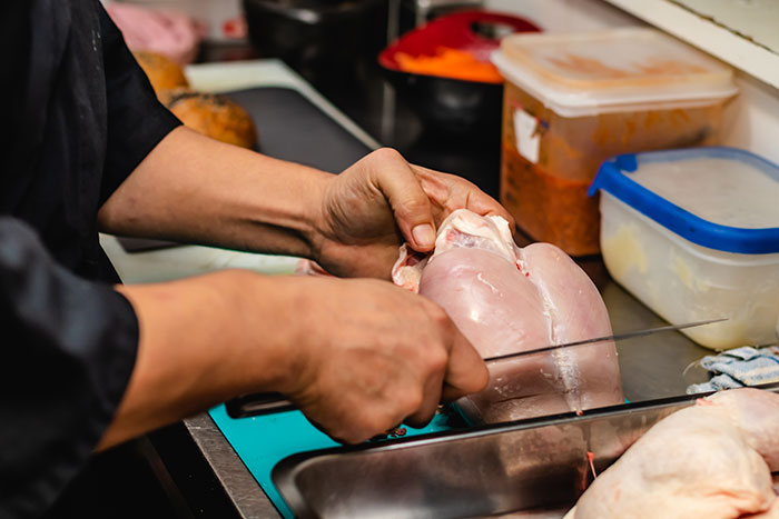 Hands slicing raw chicken breast on a cutting board in a kitchen, highlighting concerns about how chicken is prepared. Hands slicing raw chicken breast on a cutting board in a kitchen, highlighting concerns about how chicken is prepared.