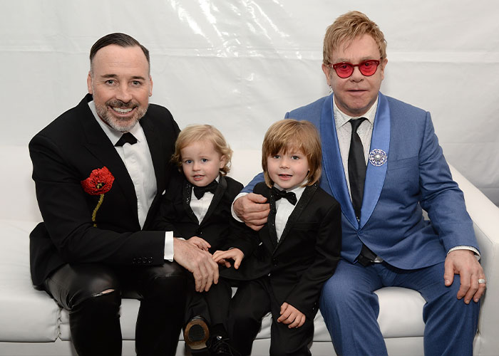 Two men and two young boys in formal suits, highlighting celebrity families and surrogacy backlashes.