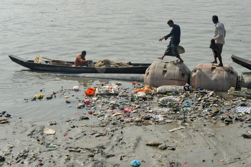 Polluted river water with floating trash and people in a boat and standing near large containers, showing dirty countries pollution.