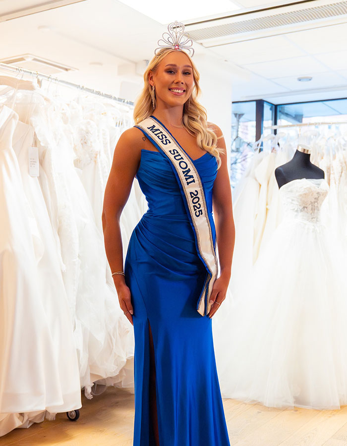 Miss Finland wearing blue gown and crown, posing indoors among wedding dresses before losing crown amid social media backlash. Miss Finland wearing blue gown and crown, posing indoors among wedding dresses before losing crown amid social media backlash.