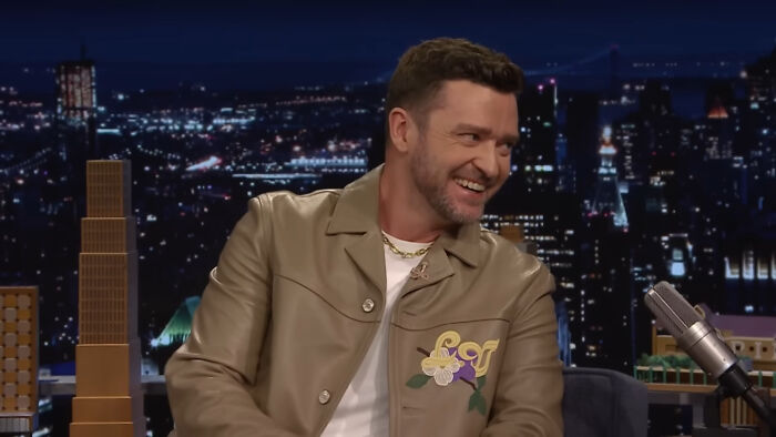 Justin Timberlake smiling on a late night talk show set, wearing a tan jacket and gold chain necklace.