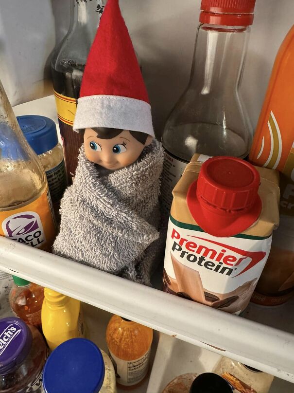 Elf on the shelf wrapped in a towel inside a fridge door among various bottles and cartons, creative mischief idea.