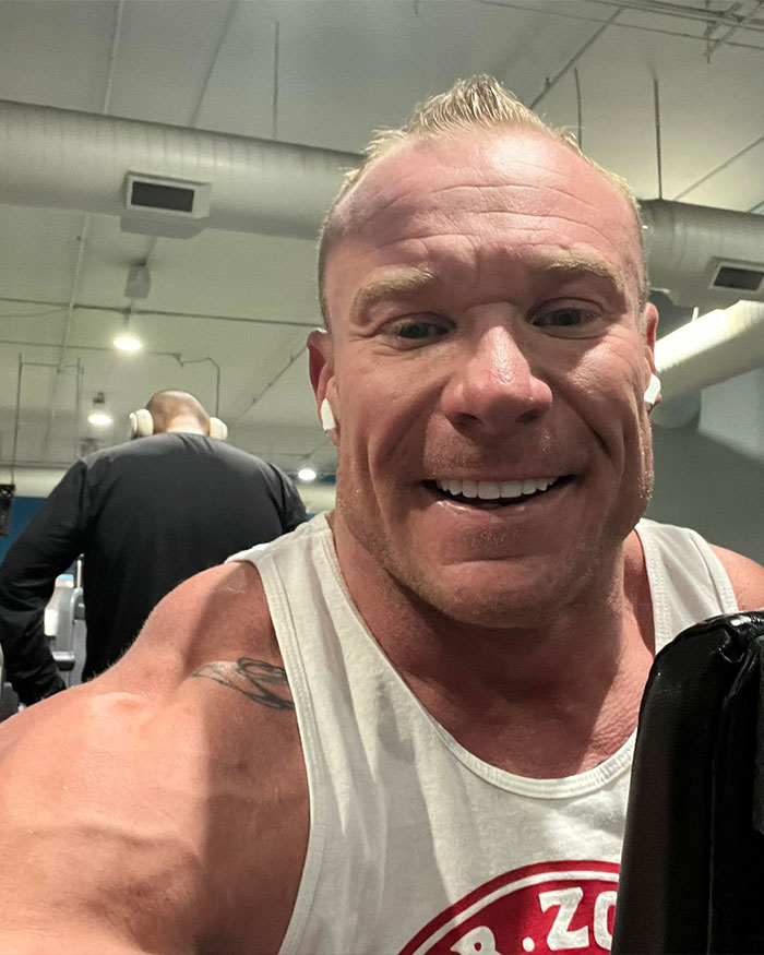 Muscular man smiling in a gym, wearing a white tank top and earbuds, with focus on his face and shoulders.