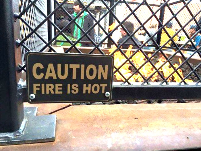 Caution sign reading fire is hot on a metal fence with people and a fire burning behind, funny memes concept.