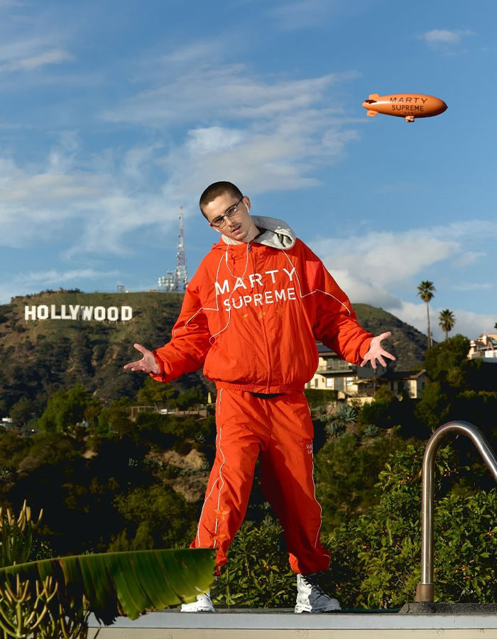 Man in red tracksuit posing near Hollywood sign under blue sky during daytime, reflecting Timoth&eacute;e Chalamet arrogance controversy.