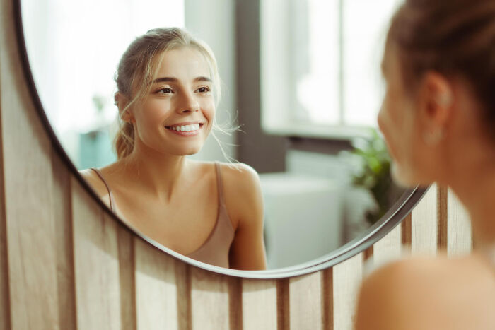 Young woman smiling at her reflection in a round mirror, capturing an embarrassing moment people were blessed to see.