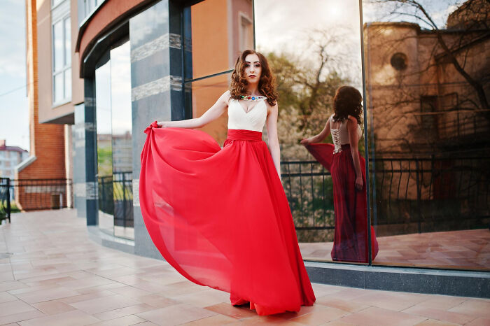 Woman in a long red dress posing near a reflective window capturing an embarrassing moment people were blessed to see.