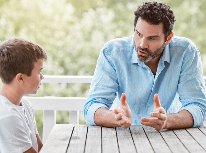 Man in blue shirt having a serious conversation with a boy at wooden table, highlighting next-level evil kids concept.