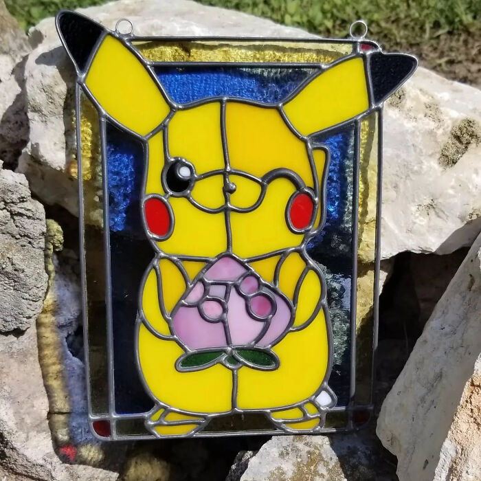 Stained glass art of a popular pop culture icon Pikachu holding a pink flower, displayed outdoors on rocks.