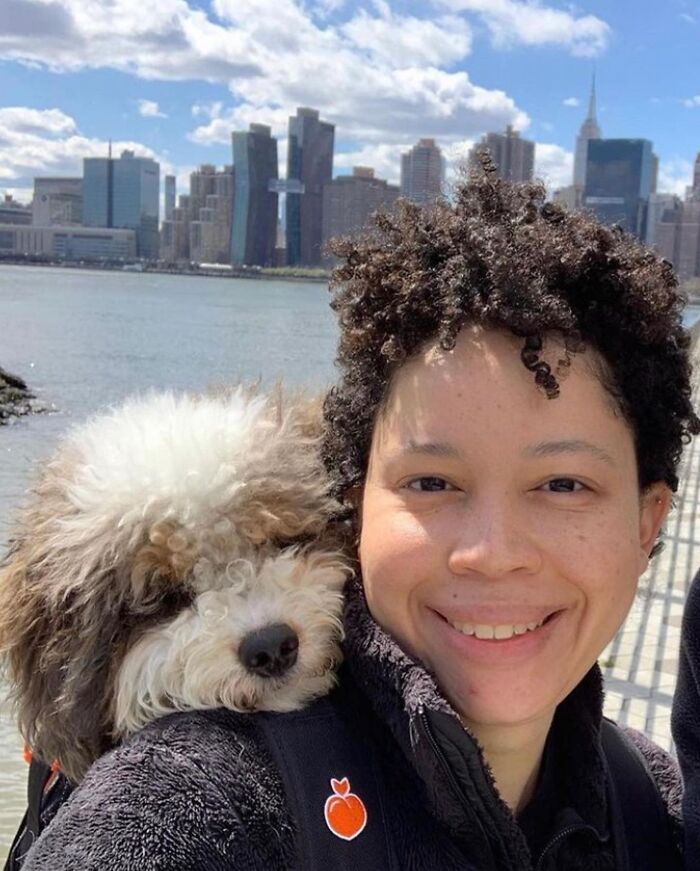 Smiling person with curly hair and a fluffy dog in a backpack, enjoying city adventures by the waterfront skyline.