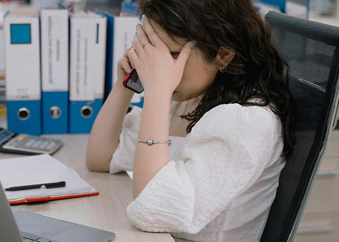 911 operator overwhelmed with a non-emergency call, holding phone and covering face in a busy office setting.