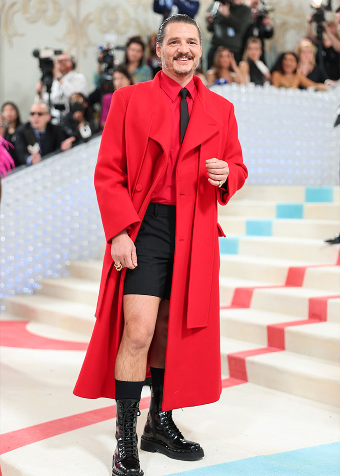 Man wearing bold red coat, black shorts, and boots walking on a red carpet highlighting celebrity fashion disasters 2025.