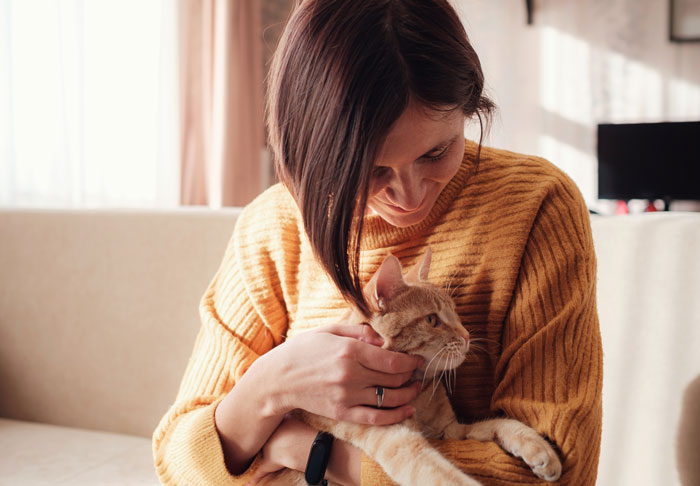 Woman in a mustard sweater holding an 11-year-old cat, relating to family conflict over cat's shared name with son. Woman in a mustard sweater holding an 11-year-old cat, relating to family conflict over cat's shared name with son.