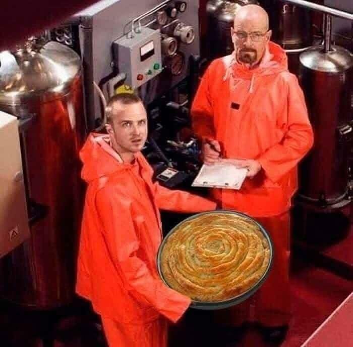 Two men in orange hazmat suits in industrial setting holding a large traditional Eastern European pastry spiral meme.