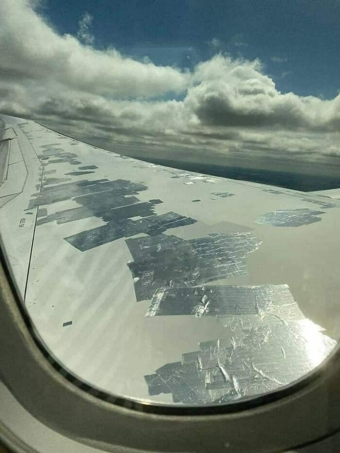 Airplane wing covered with multiple layers of silver duct tape seen through window against cloudy sky Eastern Europe meme.