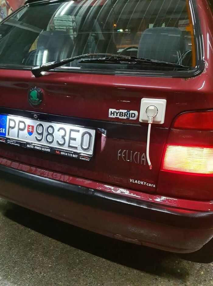 Red car with c*****d bumper and a fake hybrid badge with an electrical plug socket attached, illustrating Eastern Europe humor.