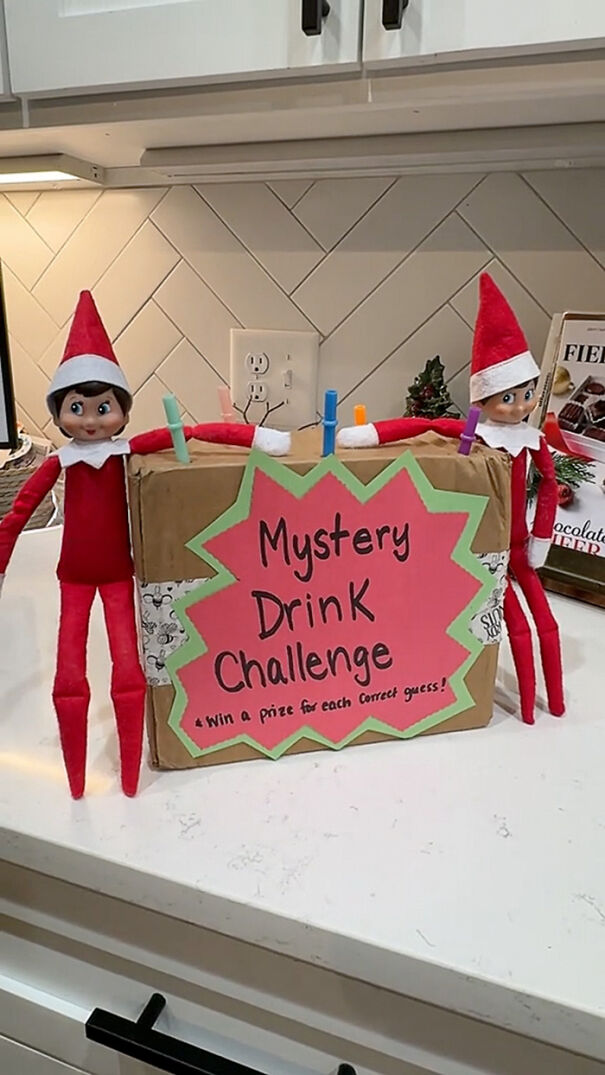 Two Elf on the Shelf dolls holding a Mystery Drink Challenge box with colorful straws in a kitchen setting.