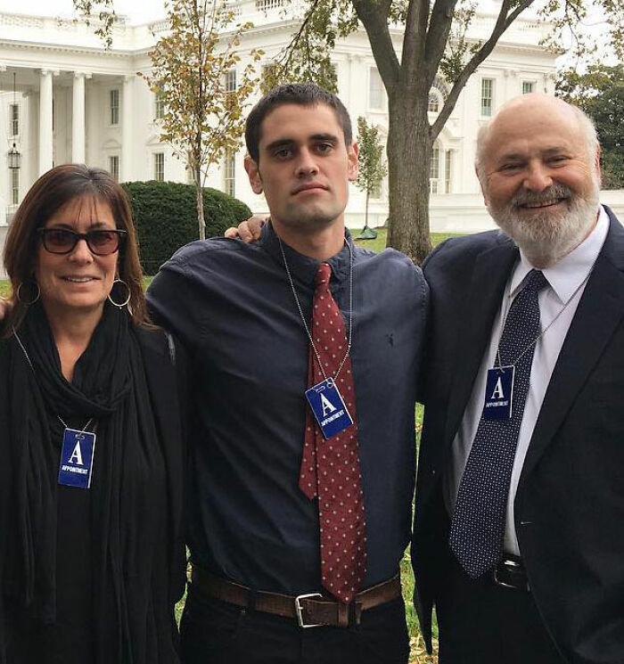 Nick Reiner standing between two people outdoors at an event, dressed formally with press badges visible on each.