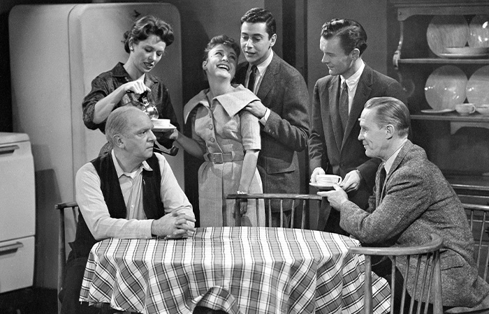Black and white scene from one of the longest running TV shows, featuring six people around a kitchen table in vintage clothing.