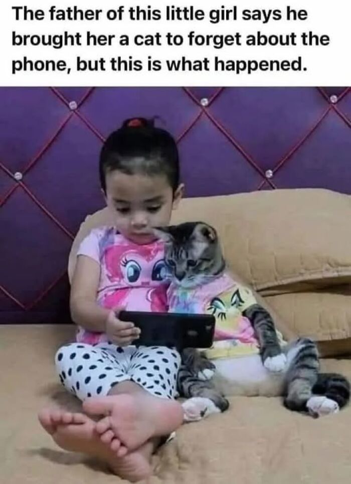 Little girl and cat wearing matching shirts sitting on a couch, cat behaving like a jerk while using a phone.
