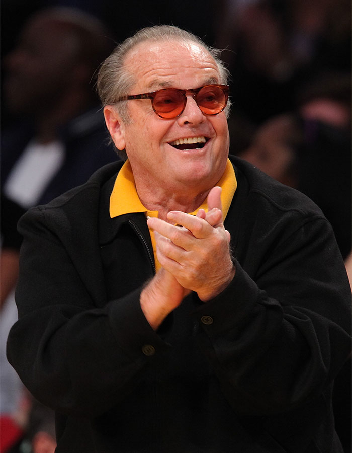 Older man wearing orange sunglasses and a black jacket clapping at an event related to outrageous actor contract demands