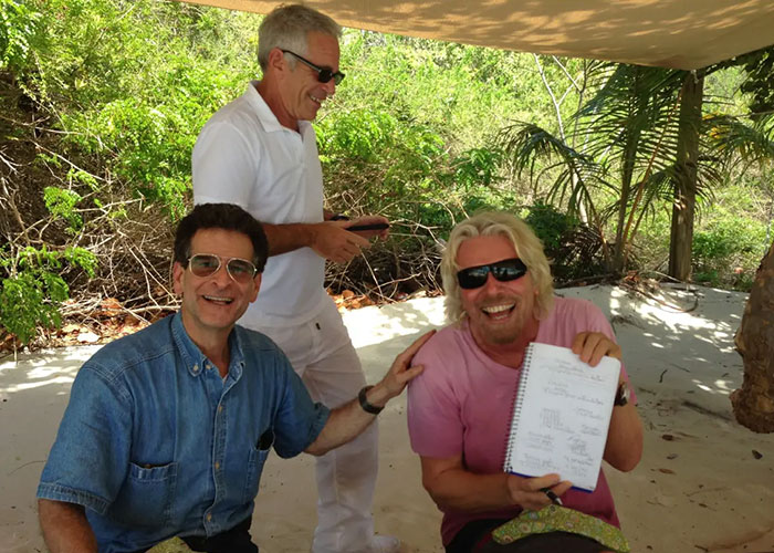 Three men outdoors, one showing a notebook, with a setting triggering backlash over disturbing Epstein desk photo.