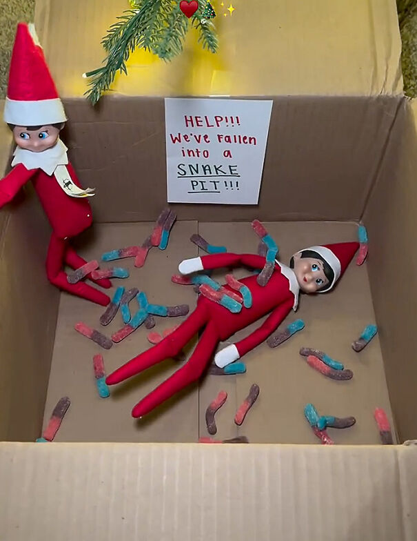 Two Elf On The Shelf dolls in a cardboard box filled with gummy worms, staged as a playful snake pit prank.